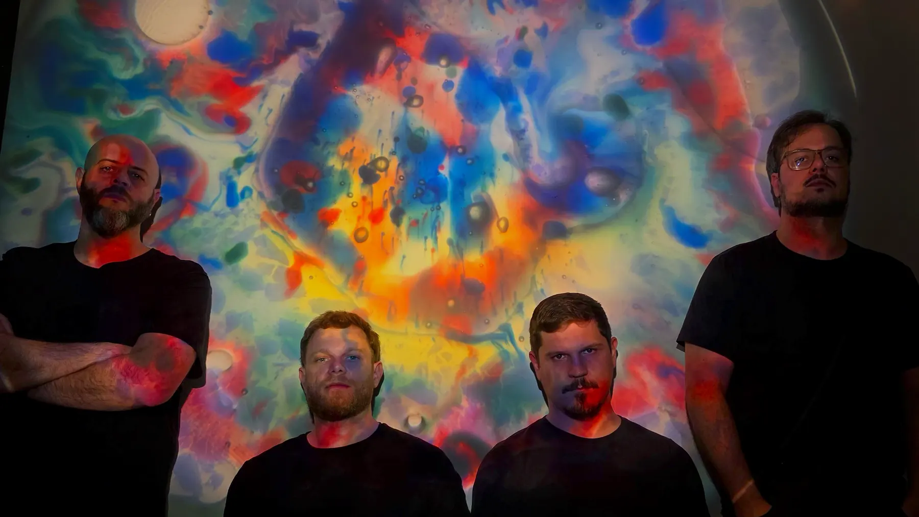 Os quatro integrantes da Birdless de camiseta preta diante de uma projeção colorida de tinta e óleo